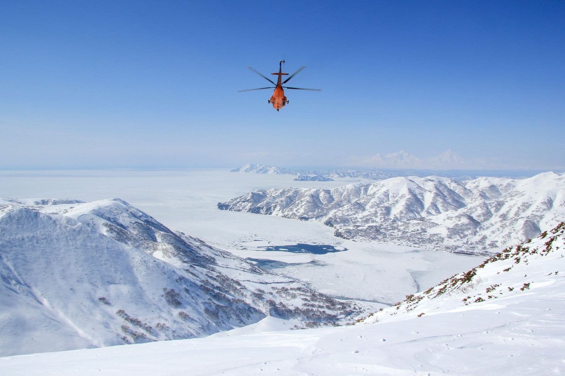 A helicopter flys down the mountain with the ocean in the background in search of lines to ski. Nobody around. Paradise. 