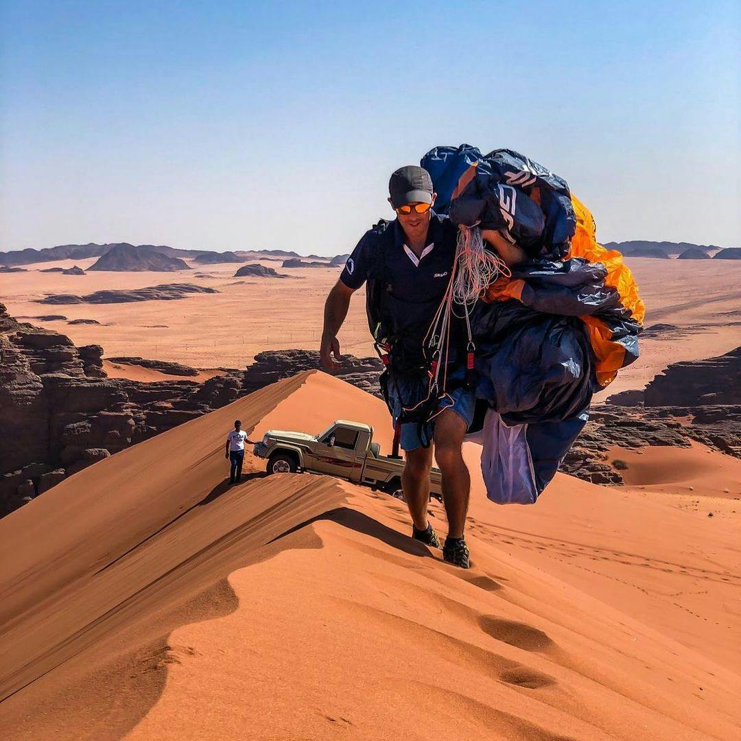 Man carrying paramotor on top of a dune in the desert with a 4x4 in the background