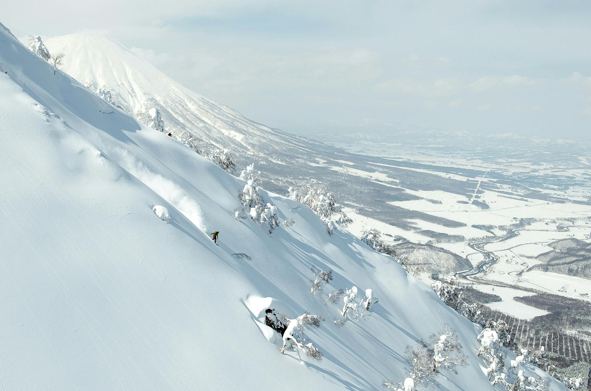 A snowboarders slides down a mountain covered with snow and a Japanese volcano observes in the background.